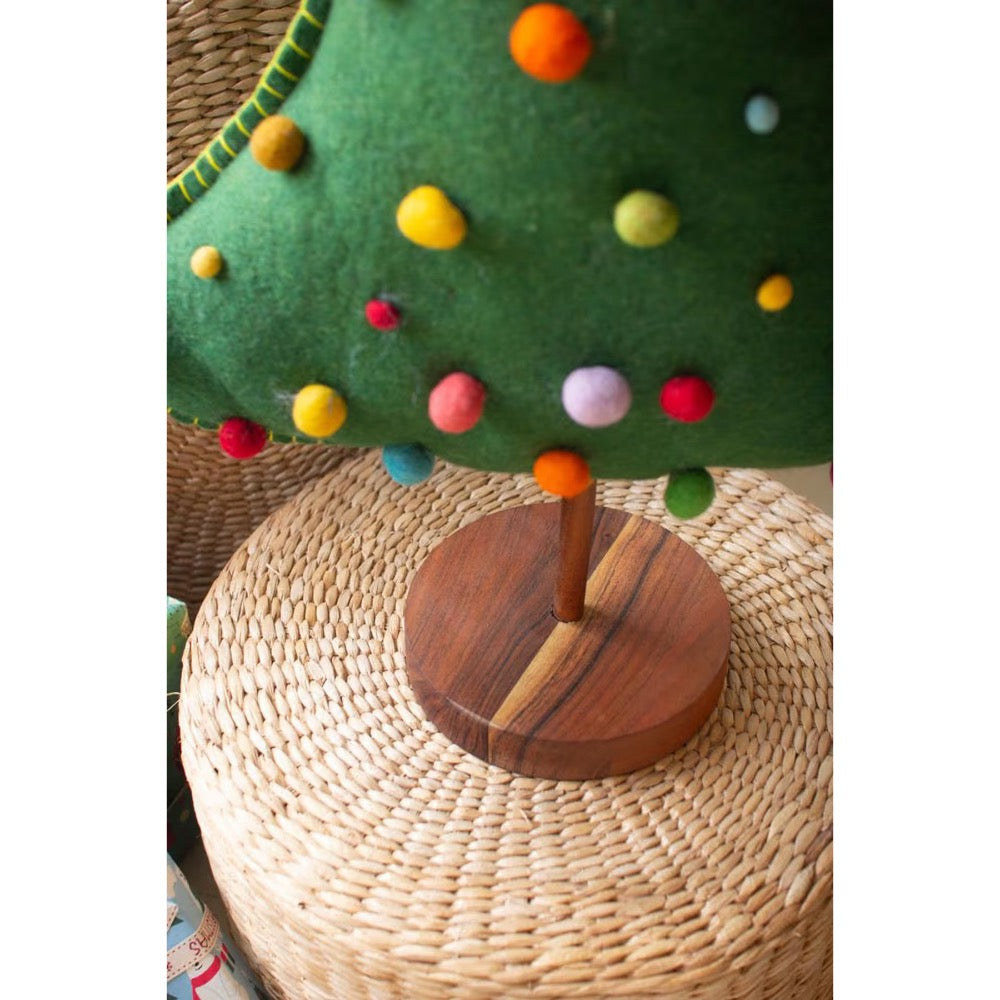 Close up for bottom part of green felt christmas tree with colored pom pom balls and a wooden base on a rattan riser.