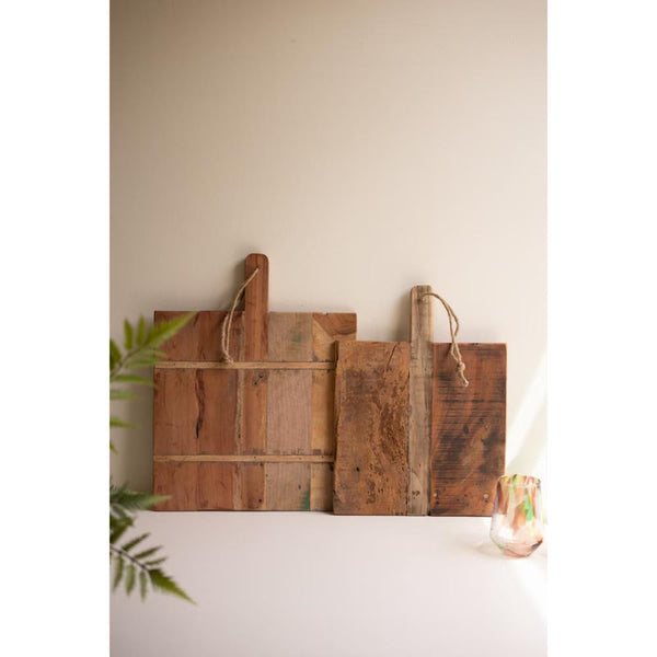 Three rustic reclaimed wood cutting boards with rope handles, arranged on a white surface against a beige wall, with a green plant leaf on the left and a small glass vase on the right.
