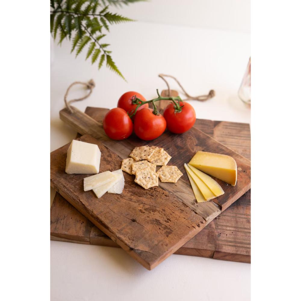 Two rustic wooden serving boards stacked on a white surface, styled with red tomatoes on the vine, crackers, and wedges of cheese. A green fern leaf and a glass bottle are softly blurred in the background.