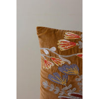 A close-up of a brown velvet throw pillow featuring detailed floral embroidery in red, yellow, and lavender with silver-gray outlines, set against a plain light gray background.
