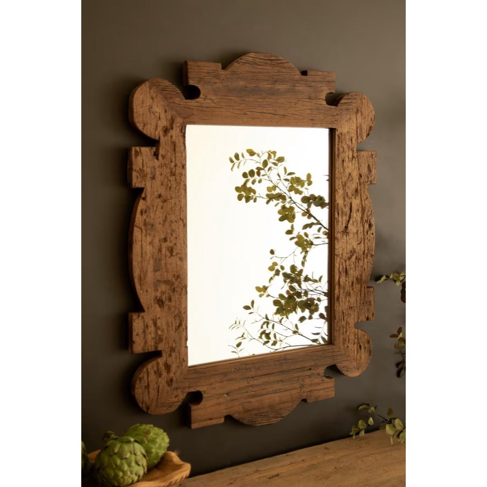 A rustic rectangular wall mirror with an ornate carved wooden frame in a medium brown finish, mounted on a dark gray wall above a wooden surface with green plant accents.