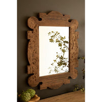 A rustic rectangular wall mirror with an ornate carved wooden frame in a medium brown finish, mounted on a dark gray wall above a wooden surface with green plant accents.