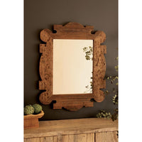 A rustic rectangular wall mirror with an ornate carved wooden frame, mounted on a dark brown wall above a wooden console table with decorative greenery.
