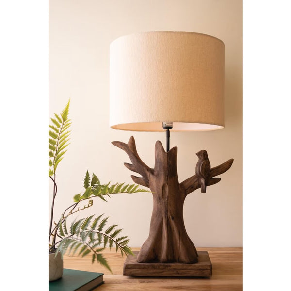 A rustic wooden table lamp shaped like a tree trunk with a small bird perched on one branch, featuring a beige fabric drum shade, displayed on a wooden surface next to a potted fern.