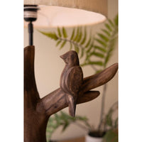 A rustic wooden bird-shaped lamp shade or decorative lamp base perched on a branch, with a warm glowing lampshade above and blurred green plants in the background.