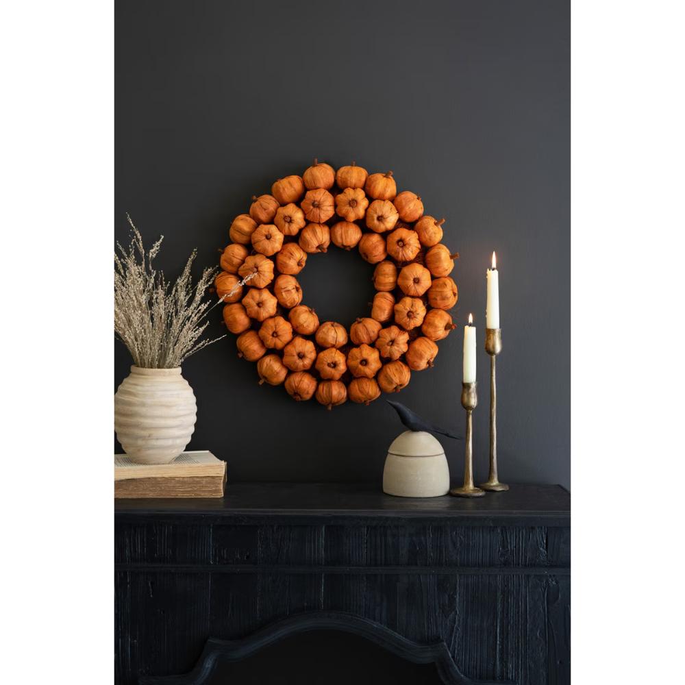 An autumn-themed wreath made of small orange pumpkins hangs on a dark charcoal wall above a black mantel decorated with a ceramic vase of dried grass, candles, and a small decorative pot.