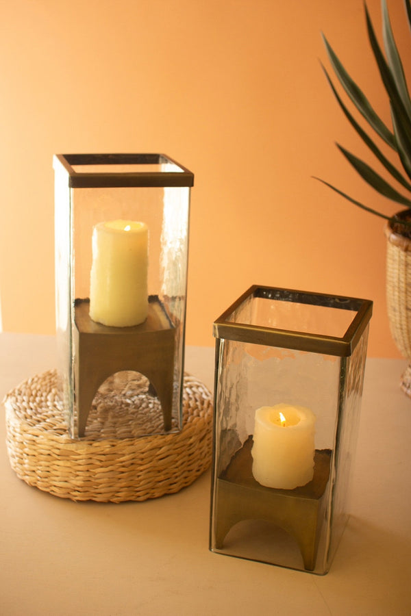 Two glass candle holders with candles on a surface against an orange wall.