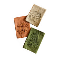 Three square linen napkins in peach, beige, and sage green, each featuring an embroidered pumpkin design, arranged on a plain white background.