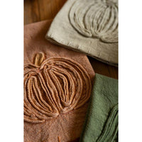 Three textured fabric napkins featuring raised pumpkin designs, shown in warm brown, beige, and sage green on a rustic wooden surface.