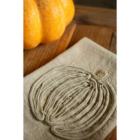 A close-up of a beige fabric napkin or towel featuring an embroidered pumpkin design, styled on a wooden surface with a real pumpkin in the background.