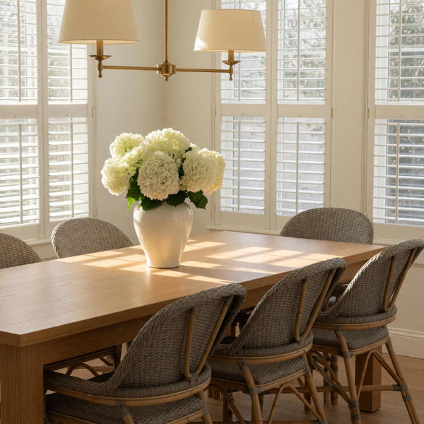 A dining room scene with a light wood rectangular table surrounded by woven gray chairs, set in front of white plantation shutters with a vase of white flowers on the table and a brass wall sconce above.