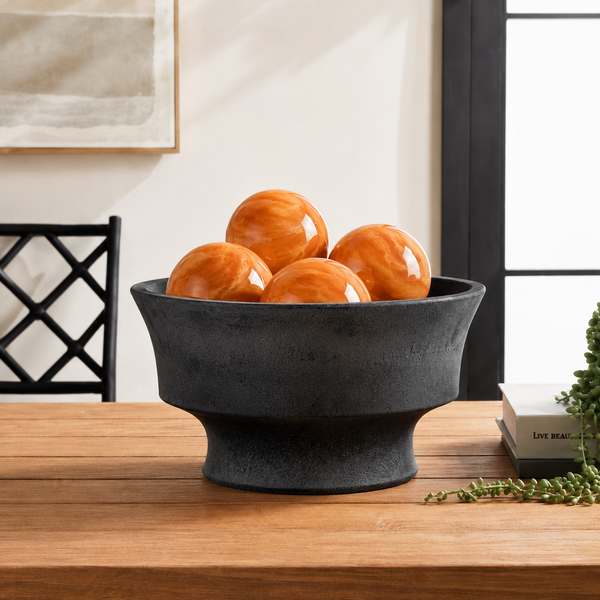 A black bowl holds five Napa Home & Garden Alani Enameled Iron Orange Orbs on a table with books and greenery.