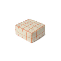 A square pouf ottoman with a cream-colored fabric upholstery and a thin orange grid plaid pattern, shown on a plain white background.