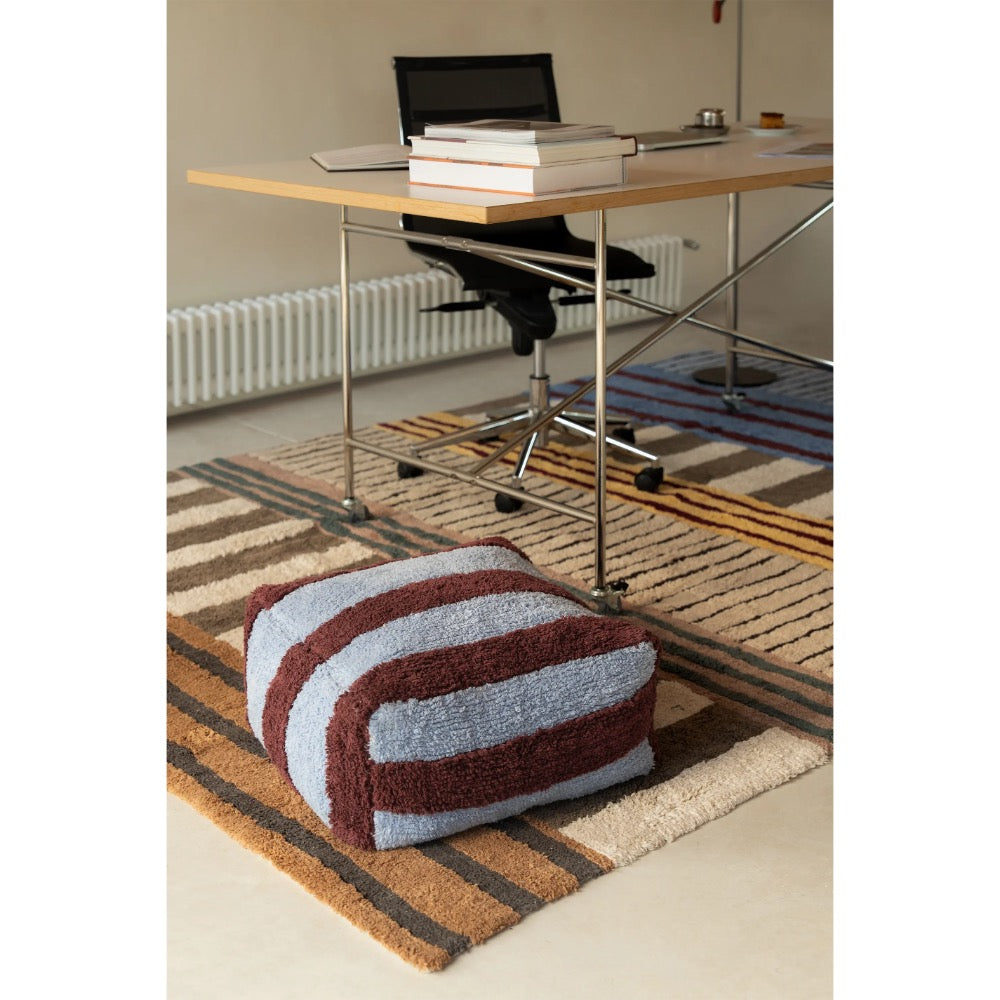 A striped pouf cushion in brown, beige, and light blue sits on a patterned rug in a modern home office setting with a desk and chair in the background.