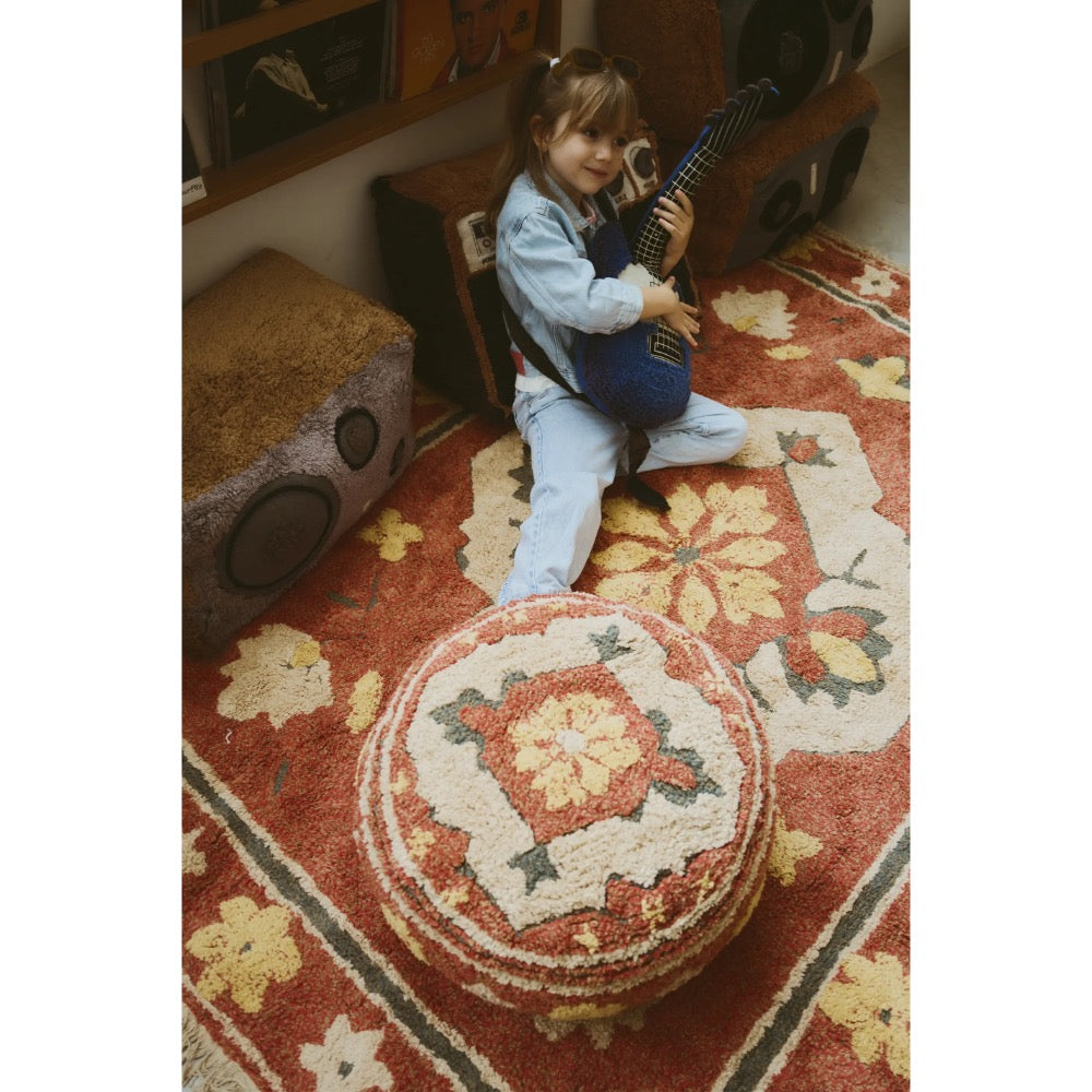 A young girl sits on a patterned red and cream rug in a cozy room, holding a blue acoustic guitar. A matching round tufted pouf sits in the foreground, with vintage-style speakers and framed art in the background.
