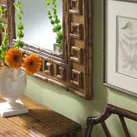 Orange flowers in a vase sit on a table by Kenian’s Tortoise Shell Bamboo Greek Key Mirror and botanical wall art.