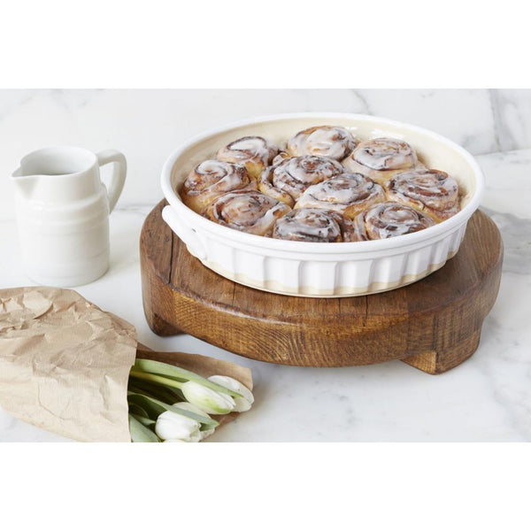 Casserole dish with cinnamon rolls on a wooden trivet, next to a pitcher and a loaf of bread on a white surface.