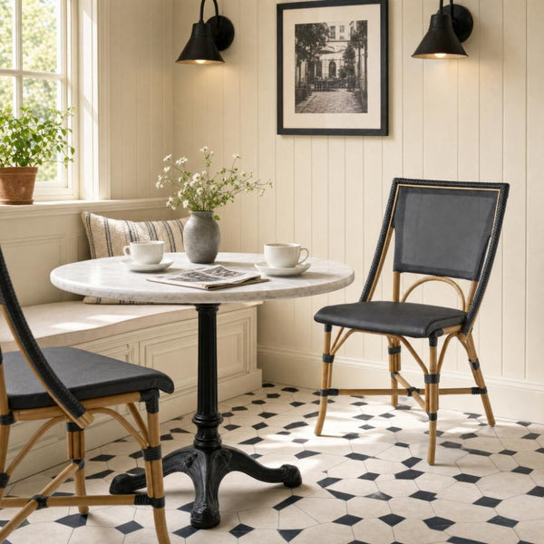 A round marble-top cafe table with a black pedestal base, surrounded by two black-and-wood dining chairs in a bright, cream-paneled room with a built-in bench, wall sconces, and a black-and-white diamond-pattern tile floor.