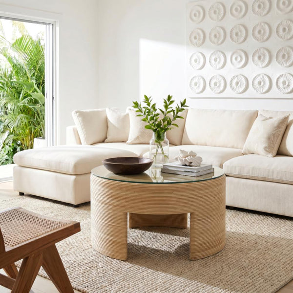 A modern living room featuring a cream sectional sofa, a round woven rattan coffee table with a glass top, a natural fiber rug, and a large white textured wall art piece.