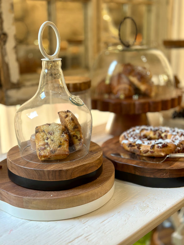 Wooden stand with a glass container holding cookies on a table with other pastries in the background.