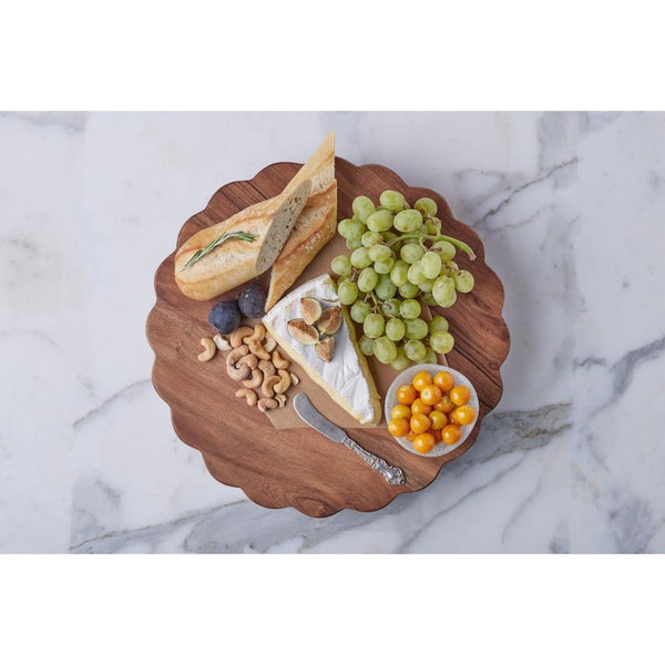 Scalloped wooden charcuterie board with bread, cheese, fruits, and nuts on a marble surface