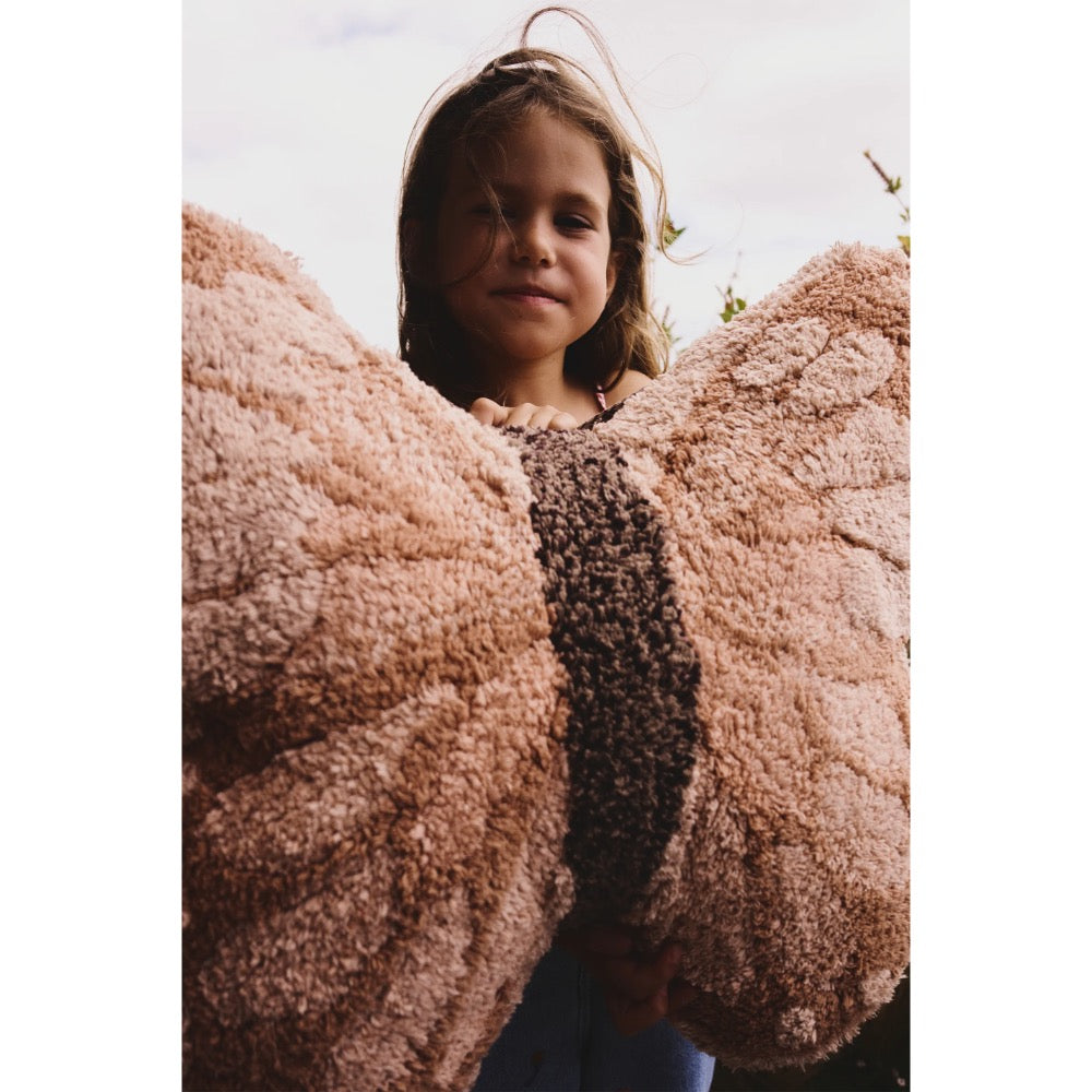 A young girl with light brown hair stands behind a large, plush, butterfly-wing-shaped throw blanket in a dusty rose pink color with a dark brown center stripe. The photo is taken from a low angle, looking up at the child, with a soft, overcast sky in the background.