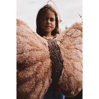 A young girl with light brown hair stands behind a large, plush, butterfly-wing-shaped throw blanket in a dusty rose pink color with a dark brown center stripe. The photo is taken from a low angle, looking up at the child, with a soft, overcast sky in the background.