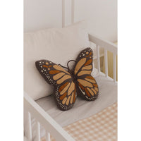 A butterfly-shaped decorative pillow with orange, brown, and cream patterned wings rests on a white crib next to a white pillow.