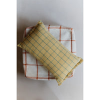 A rectangular throw pillow with a mustard yellow and teal plaid pattern and fringed edges, resting diagonally on top of a white and rust plaid blanket against a neutral light gray background.