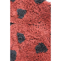 Close-up of a shaggy rug with a coral red base and irregular dark gray spots, shown against a white background.