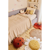 A cozy children's bedroom scene with a single bed dressed in beige bedding, a gray upholstered headboard, decorative pillows, and plush floor cushions shaped like a ladybug, flower, and sun on a light wood floor.
