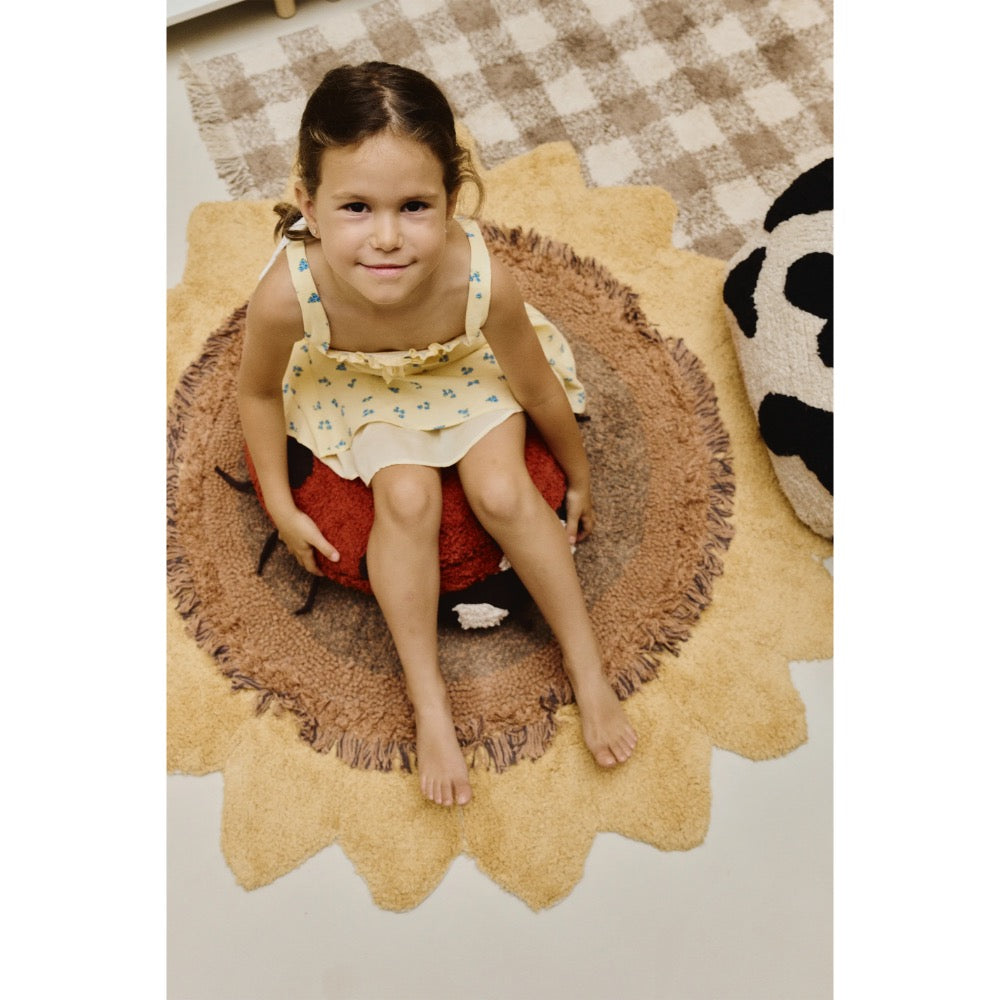A young girl sits barefoot on a round, scalloped beige rug with a brown fringe, positioned over a red and black pumpkin-shaped cushion. The room features a brown and beige checkered rug on the floor.