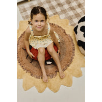 A young girl sits barefoot on a round, scalloped beige rug with a brown fringe, positioned over a red and black pumpkin-shaped cushion. The room features a brown and beige checkered rug on the floor.