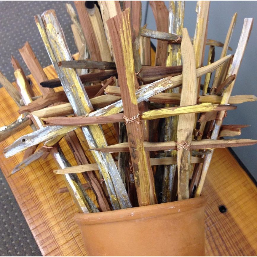The Gold + Silver Rustic Cross in Frame - Large by Shadow Catchers Art complements the wood sticks, artfully woven in a clay pot on a wooden surface.