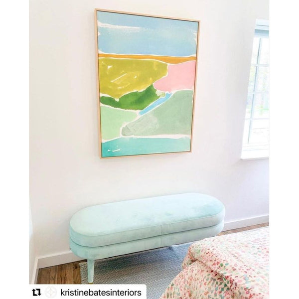 The Watercolor Framed Art - Abstract Italian Landscape I by Shadow Catchers Art, featuring a pastel abstract design reminiscent of the optic pop style, is gracefully displayed on the wall above a light blue cushioned bench in a bright room.