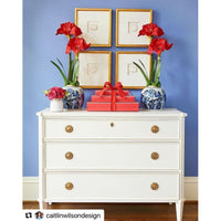 A white dresser embellished with red gifts and flowers, along with blue vases and four framed art pieces on a blue wall, features one piece showcasing the Framed Intaglio Art in Gold - Style 1 from Shadow Catchers Art.