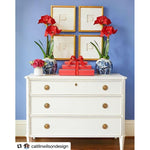 A white dresser adorned with red flowers in blue vases, stacked gifts, and four square frames featuring engraved intaglio images from Shadow Catchers Art's Framed Intaglio Art in Gold - Style 8 on a blue wall.