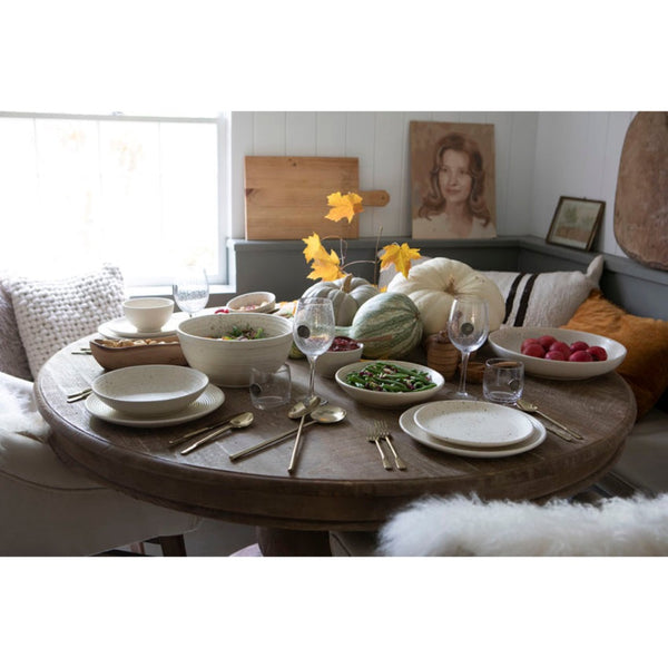 A round wooden dining table set for a meal in a bright, modern dining room. The table is styled with white ceramic plates, bowls, gold-toned cutlery, wine glasses, and autumn decorations including pumpkins, a green melon, and yellow artificial leaves.