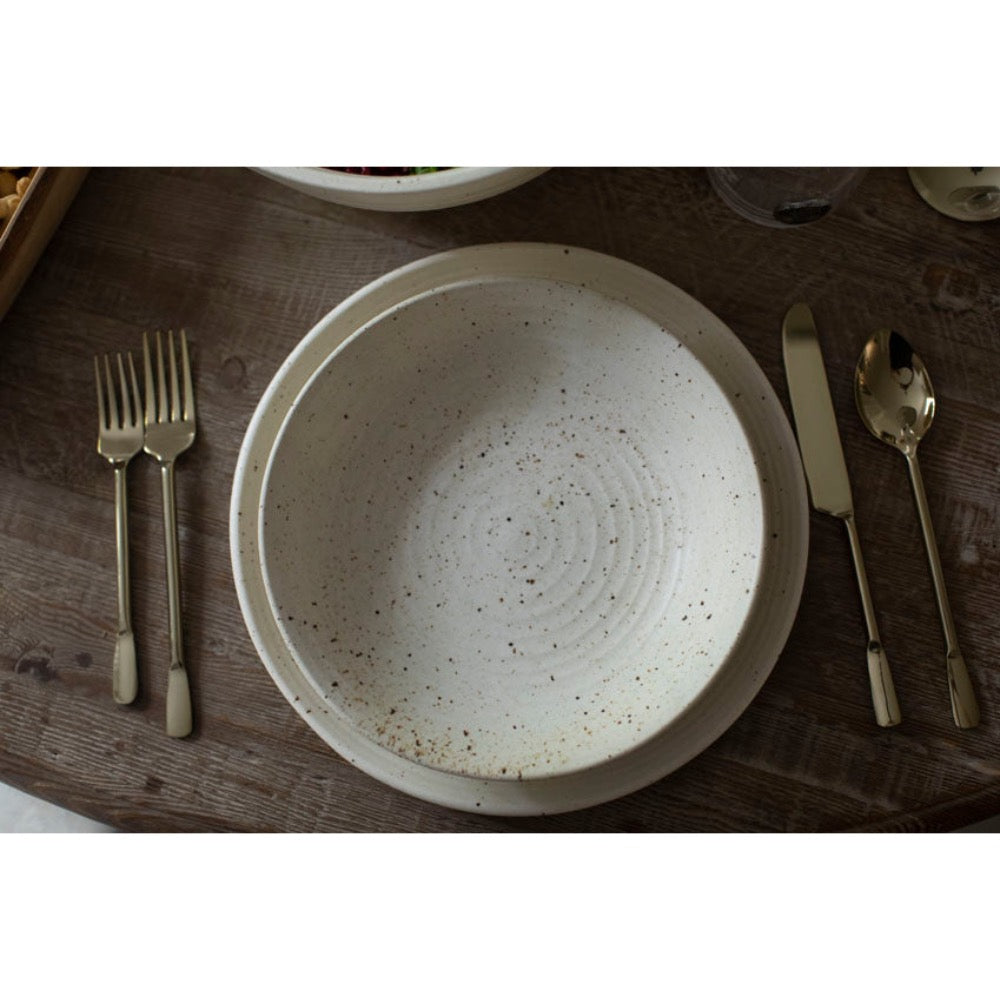 A round speckled cream-colored ceramic bowl with concentric ring patterns, styled on a dark wooden table with gold-toned cutlery.