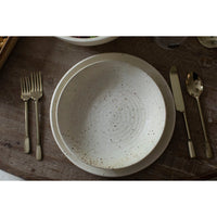 A round speckled cream-colored ceramic bowl with concentric ring patterns, styled on a dark wooden table with gold-toned cutlery.