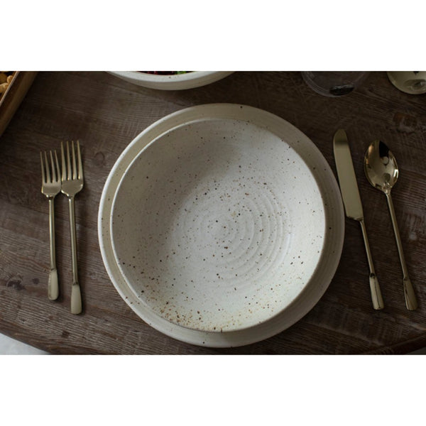 A round speckled cream-colored ceramic bowl with concentric ring patterns, styled on a dark wooden table with gold-toned cutlery.