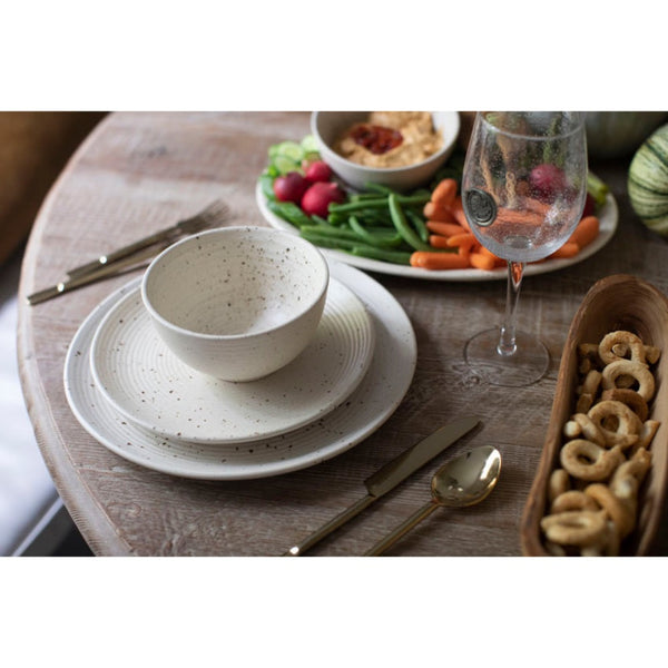 A rustic ceramic dinnerware set on a round wooden dining table, featuring a speckled cream-colored bowl, matching saucer, and coordinating plates, styled with gold-toned cutlery, a glass of water, and fresh vegetables.