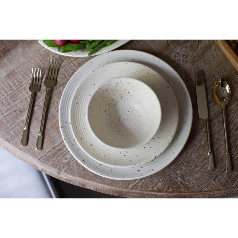 A ceramic dinnerware set featuring a speckled cream-colored plate and matching bowl, styled on a rustic wooden table with silver and gold flatware.