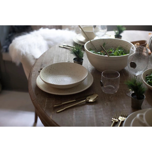 A styled dining table setting featuring speckled beige ceramic plates, a bowl of salad, clear drinking glasses, and gold-toned cutlery on a rustic wooden table.