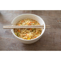 A ceramic bowl filled with stir-fried noodles and vegetables, with wooden chopsticks resting across the rim, set on a rustic wooden tabletop.