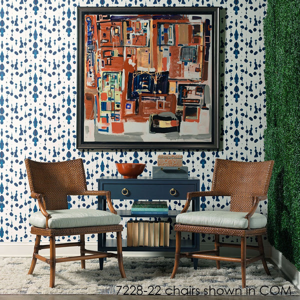 A styled interior scene showing two woven wicker armchairs with pale green cushions placed on a rug in front of a blue console table, with a large framed abstract painting on a blue-and-white patterned wallpaper wall.