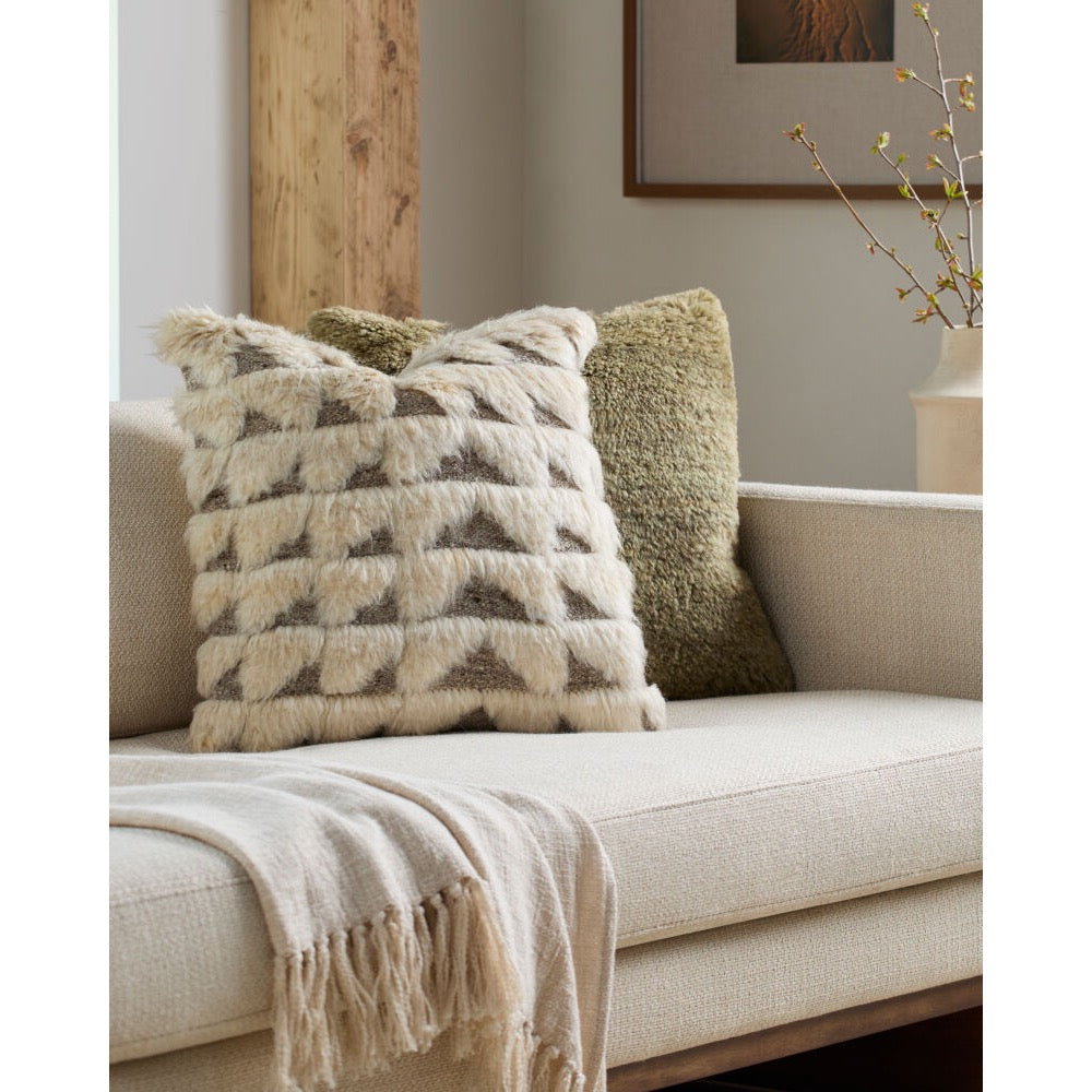 A textured cream throw pillow with a geometric pattern sits on a beige sofa next to an olive green shag pillow and a cream throw blanket in a styled living room setting.