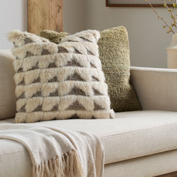 A textured cream throw pillow with a geometric pattern sits on a beige sofa next to an olive green shag pillow and a cream throw blanket in a styled living room setting.