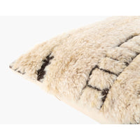 Close-up of a fluffy sheepskin or wool throw pillow in cream with dark brown geometric woven patterns, shown on a plain white background.