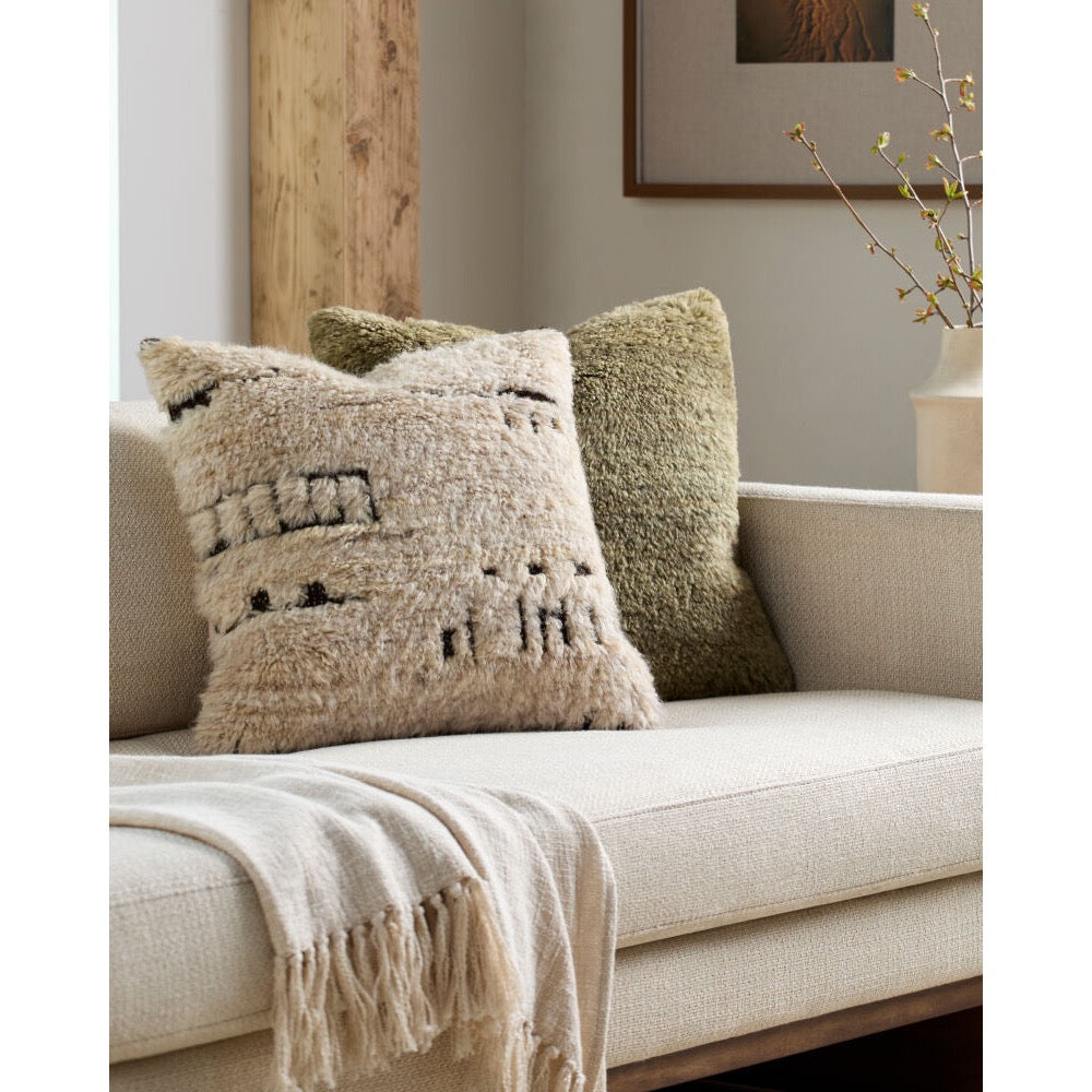 Two textured throw pillows on a light beige sofa in a styled living room setting. The front pillow is cream with black geometric woven patterns, and the back pillow is olive green.